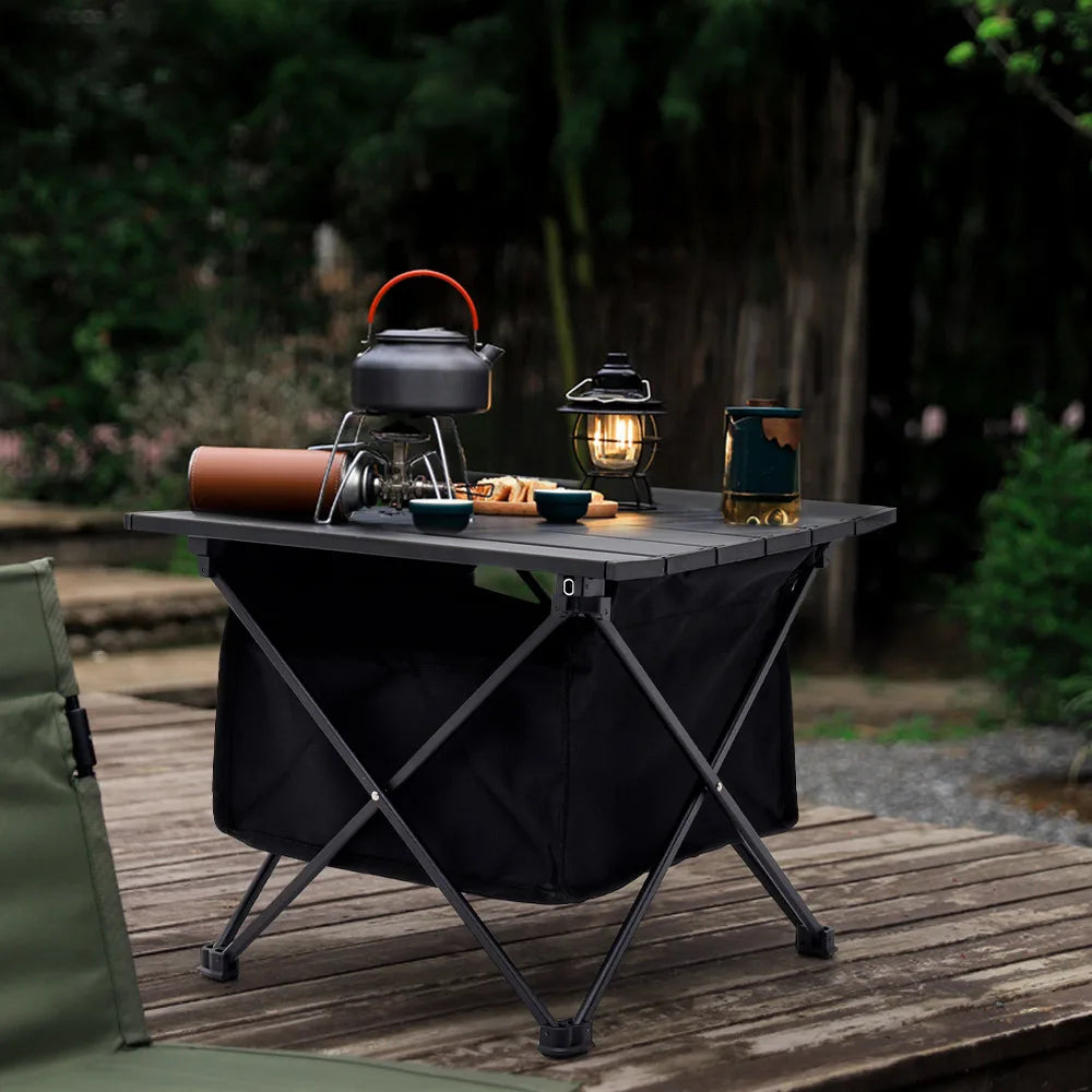 Black folding camping table on wooden deck with slatted top and storage bin